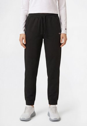 Persona che indossa pantaloni jogger neri con coulisse, tasche laterali e scarpe da ginnastica bianche, in piedi davanti a uno sfondo bianco uniforme.