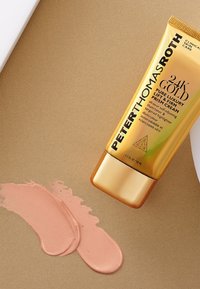 Guldrör med 24K Gold Pure Luxury Lift & Firm Prism Cream med svart text. Två rosa krämprover är utsmetade på en gyllene yta.