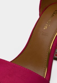 Κοντινή λήψη ροζ σουέτ γόβας με ψηλό τακούνι, καφέ πάτο με χρυσές λεπτομέρειες στην άκρη και ανάγλυφο λογότυπο της μάρκας.