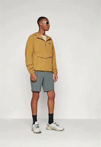 Chaqueta de cremallera marrón claro con un bolsillo frontal, combinada con pantalones cortos grises. El modelo lleva gafas de sol blancas y zapatillas deportivas de color gris claro.