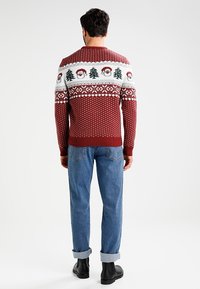 Sweter wełniany w kolorze bordowym, z białym i zielonym wzorem bożonarodzeniowym przedstawiającym Świętego Mikołaja i choinki. Ściągacze na mankietach i u dołu. Zestawiony z niebieskimi dżinsami.