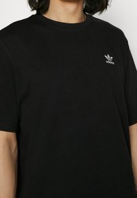 Černé bavlněné tričko s kulatým výstřihem, krátkými rukávy a malým bílým logem Adidas na levé části hrudi. Měkká textura.