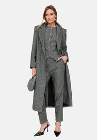 Grauer Hahnentritt-Anzug mit maßgeschneidertem Blazer und taillierter Hose, ausgestattet mit goldenen Knöpfen. Kombiniert mit schwarzen High Heels und einer grauen Handtasche.