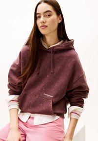 Sweat à capuche marron foncé avec une texture usée, poche avant et capuche à cordon. Porté sur une chemise blanche à col et accompagné d'un pantalon rose.