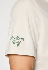 Kortermet beige polo-shirt med grønn brodert "Malbon Golf"-logo på venstre erme. Mykt stoff med en glatt tekstur.