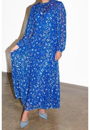 Femme portant une longue robe bleue à motif cachemire avec des manches transparentes et des chaussures pointues bleu clair avec des boucles décoratives.