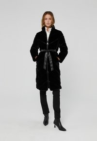 Cappotto in pelliccia sintetica nera con colletto alto, chiusura a zip, vita con cintura in tessuto satinato e motivo a quadri diagonali. Abbinato a pantaloni neri e stivali.