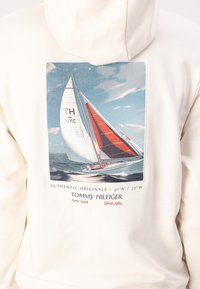 Kremfarget hettegenser med grafikk av en seilbåt med røde og hvite seil, teksten inkluderer "Tommy Hilfiger" og koordinater, glatt bomullsstoff.