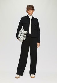 Veste noire avec poches sur un col roulé blanc, associée à un pantalon noir et des baskets beiges. Tient un sac à motifs avec des formes géométriques.
