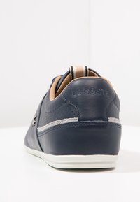 Zapatilla Lacoste azul marino con interior beige, suela blanca, costados perforados y una pestaña de tirón beige y negra en el talón.