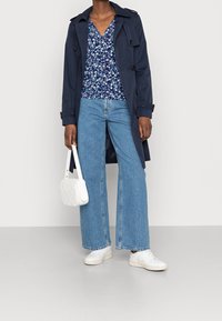 Kvinna i marinblå trenchcoat, blåblommig topp, vida jeans, vita sneakers och håller en vit handväska mot en enfärgad bakgrund.