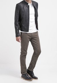 Schwarze Lederjacke mit Stehkragen, weißes Poloshirt, khakifarbene Hose und schwarze Schnürschuhe mit hellbraunen Sohlen, stehend vor einem weißen Hintergrund.