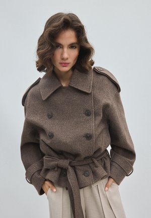 Femme aux cheveux courts et ondulés portant un manteau en laine marron croisé noué à la taille et un pantalon beige plissé sur un fond uni.