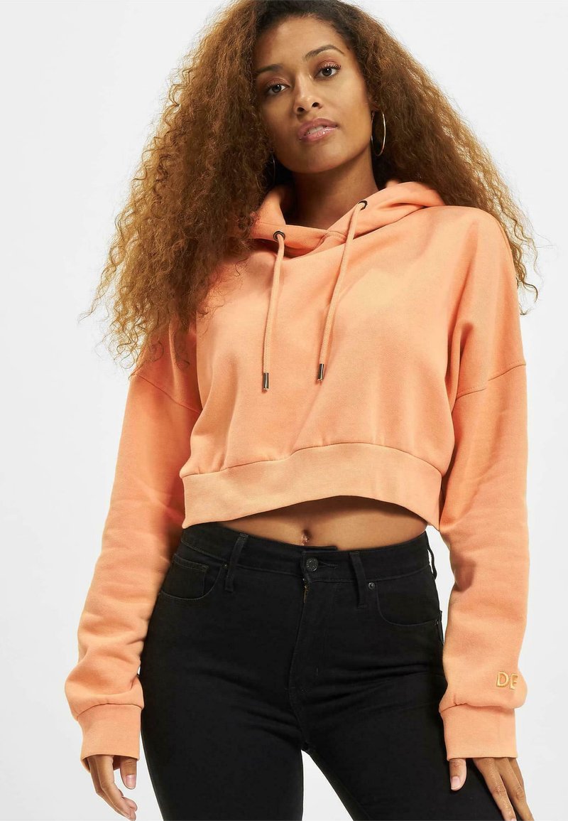 Sudadera recortada de tela color durazno con capucha ajustable. Cuenta con mangas largas y un pequeño detalle bordado en el puño.