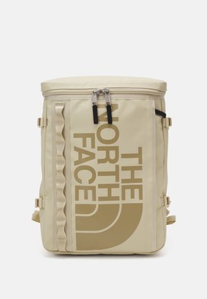 Zaino rettangolare beige con cerniera superiore, cinturini laterali di compressione e grande logo verticale "THE NORTH FACE" color tan sul pannello frontale.
