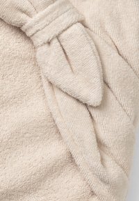 Beige frottétyg med en knuten detalj. Texturen är mjuk och plufsig, med fina öglor synliga över hela materialet.