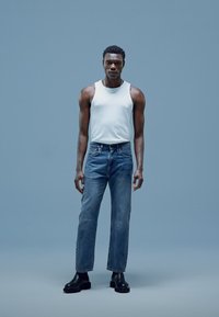 Camiseta sin mangas blanca, vaqueros rectos azul oscuro, botines negros; la modelo posa frente a un fondo azul claro.