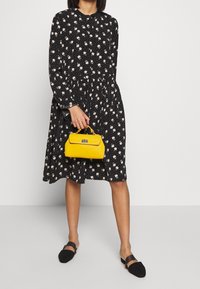 Svart blommig klänning med långa ärmar och midjesöm, kombinerad med en liten gul handväska med en strukturerad design och kedjehandtag.