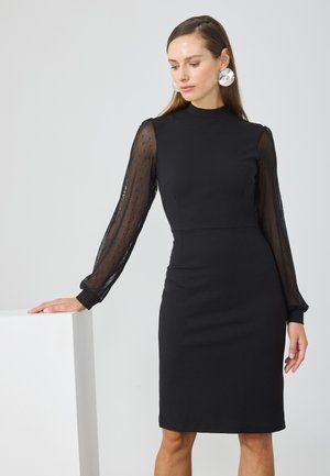 Svart tettsittende kjole med høy hals og buste design. Gjennomsiktige polka dot lange ermer med mansjett detaljer. Glatt og slett tekstur gjennom hele.