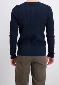 Navyblå stickad tröja med ett strukturerat mönster, rund halsringning och långa ärmar, kombinerad med ljusbruna byxor.