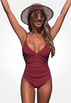 Costume da bagno intero bordeaux con scollatura profonda, texture arricciata e spalline sottili. Il modello indossa un cappello a falda larga e occhiali da sole rotondi.