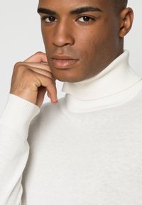 Weißer gestrickter Rollkragenpullover mit gerippten Bündchen, weicher Textur und taillierter Passform, getragen von einer Person mit neutralem Gesichtsausdruck.