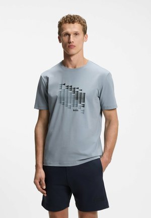Mężczyzna w jasno-niebieskim t-shircie z krótkim rękawem z abstrakcyjnym czarnym wzorem trójkątów i czarnych szortach, stoi z jedną ręką w kieszeni.