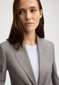 Grijze blazer met een geruit patroon en gestructureerde revers, gecombineerd met een wit T-shirt. Zachte stofstructuur met nauwkeurige stikdetails.