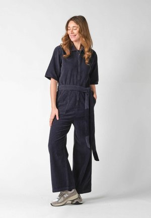 Kvinde med langt hår iført en mørkeblå fløjlsdragt med bælte, står med hænderne afslappet, smilende og ser ned.