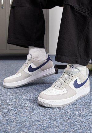 Persona che indossa pantaloni neri e calzini bianchi con scarpe Nike beige, bianche e blu navy su un pavimento con moquette blu al chiuso.