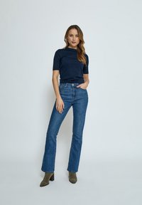 Marinblå kortärmad topp i kombination med högmidjade blå vida jeans. Modellen bär olivgröna ankelstövlar. Slät textur och figurnära design.