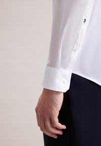 Witte shirtmouw met een knoopmanchet, voorzien van een kleine zwarte rozenbroderie. Glad stof; gecombineerd met donkerblauwe broeken.