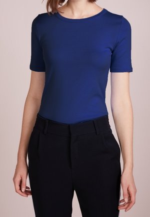 Vrouw draagt een aansluitend koningsblauw topje met korte mouwen, ingestopt in een hoge taille zwarte broek, tegen een effen achtergrond.