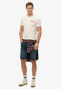 Un atuendo informal que incluye una camiseta crema con estampado rojo, shorts de mezclilla azules y una camisa de cuadros atada a la cintura. Zapatillas tostadas completan el conjunto.
