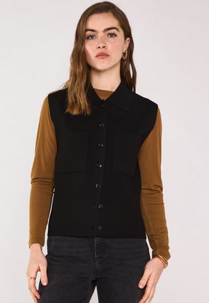 Jonge vrouw met lang bruin haar, goudkleurige creolen, een zwarte mouwloze vest met knopen, een bruin shirt met lange mouwen en een donkere spijkerbroek.