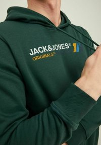 Osoba v tmavě zelené mikině s kapucí s nápisem "JACK & JONES ORIGINALS" a multibarevným logem, držící jeden šňůrku mikiny u krku.