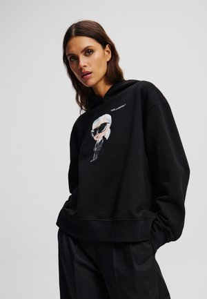 Black hoodie made of soft fabric, featuring a cartoon graphic of a character with white hair and sunglasses. Ribbed cuffs and hem.