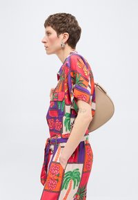 Γυναίκα με κοντά μαλλιά που φοράει πολύχρωμη στολή με λουλουδάτο και φοινικόδενδρο μοτίβο, στέκεται σε προφίλ κρατώντας μια μπεζ τσάντα ώμου.