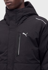 Gros plan d'une personne portant une veste noire Puma avec capuche, poche zippée et logo Puma blanc sur la poitrine.