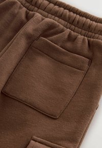 Brown sweatpants featuring a soft texture, elastic waistband, and a single back pocket with a rectangular shape.