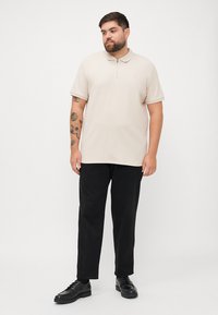 Beige short-sleeve polo shirt with a zip collar, paired with black trousers and black lace-up shoes, featuring a smooth texture and minimal design.