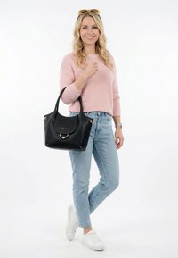 Lyshåret kvinde i lyserød sweater og jeans bærer en sort håndtaske, har hvide sneakers og solbriller på hovedet, smiler til kameraet.