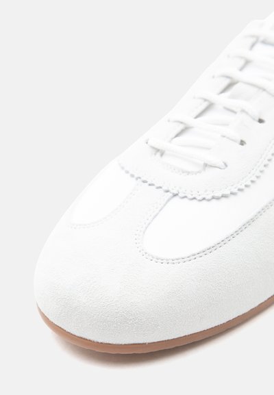 Bout de chaussure en daim blanc avec surpiqûres détaillées, bord festonné près des lacets, et semelle en caoutchouc marron clair.