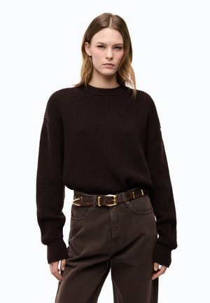 Giovane donna con capelli lisci castano chiaro che indossa un maglione di lana marrone scuro largo e pantaloni marrone scuro con una cintura fantasia.