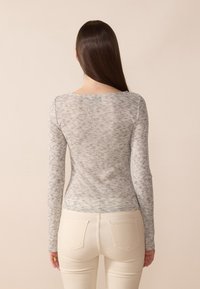 Top cu mâneci lungi, ajustat, de culoare gri deschis, cu un model texturat și melanjat. Are un decolteu larg și este asortat cu pantaloni bej ajustați.