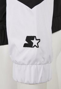 Manchon en tissu noir et blanc avec un poignet élastique, arborant un logo brodé noir en forme de "S" et une étoile.