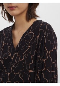 Blouse noire à encolure en V, présentant une silhouette fluide. Elle est ornée d'un motif de lignes ondulées marron et possède une texture douce et lisse.
