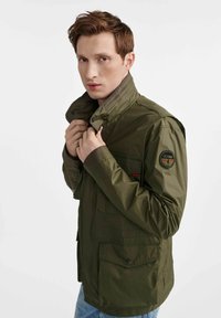 Wodoodporna kurtka w oliwkowym kolorze z wysokim kołnierzem, zapinana na zamek, z kieszenią na piersi oraz naszywką na lewym rękawie. Gładka faktura.