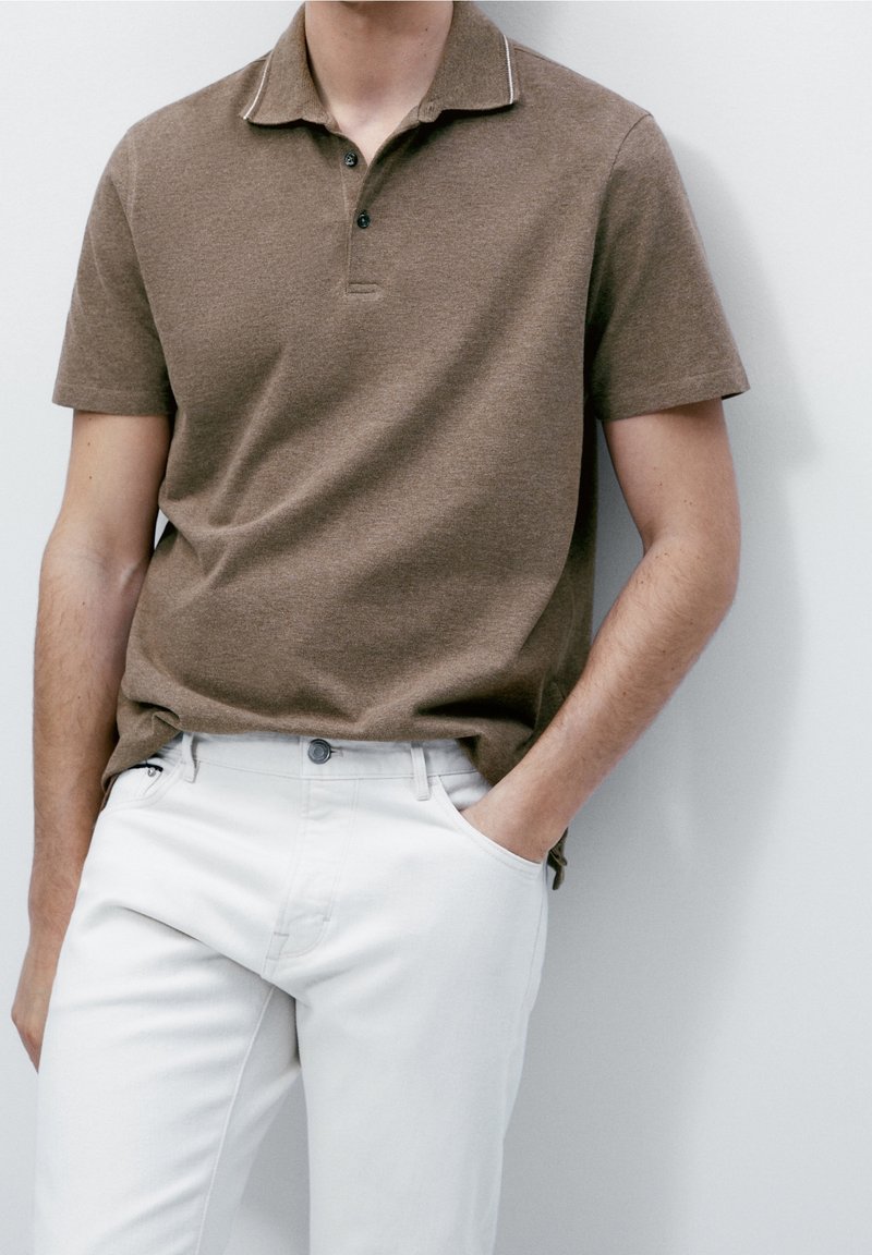Polo shirt marron avec un col et trois boutons, associé à un pantalon blanc. Tissu texturé, manches courtes et col en contraste subtil.