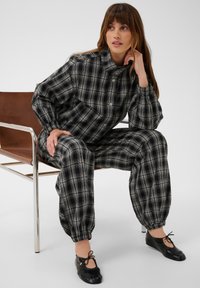 Femme portant une tenue à carreaux noire et grise boutonnée avec des poignets élastiques, assise sur une chaise en cuir marron et métal, regardant sur le côté.